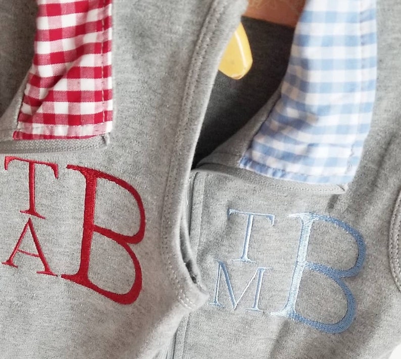 May include: Two gray sweatshirts with a zipper closure. One sweatshirt has a red and white gingham collar and the embroidered letters "T A B". The other sweatshirt has a blue and white gingham collar and the embroidered letters "T M B".