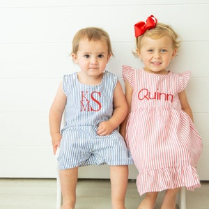 May include: Two children wearing matching red and blue striped outfits. The boy is wearing a blue and white striped romper with the monogram "KMS" embroidered on the front. The girl is wearing a red and white striped dress with the name "Quinn" embroidered on the front.