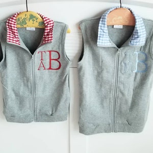 May include: Two gray fleece vests with zipper closures. One vest has a red and white checkered collar with the letters "T.A.B." embroidered in red. The other vest has a blue and white checkered collar with the letters "T.M.B." embroidered in blue.