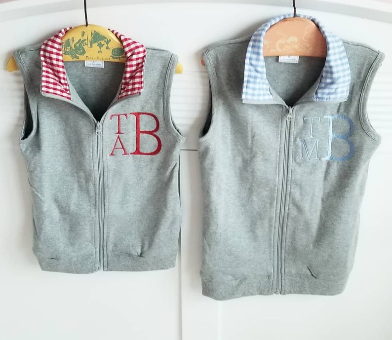 May include: Two gray fleece vests with zipper closures. One vest has a red and white checkered collar with the letters "T.A.B." embroidered in red. The other vest has a blue and white checkered collar with the letters "T.M.B." embroidered in blue.