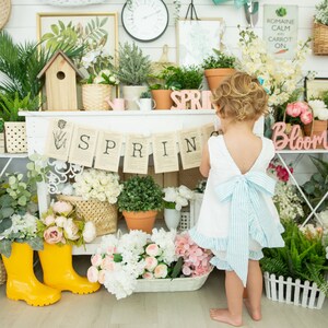 Puede incluir: Una ni&ntilde;a peque&ntilde;a con un vestido blanco y un lazo azul se para frente a un estante blanco decorado con flores, plantas y una pancarta que dice "SPRING".