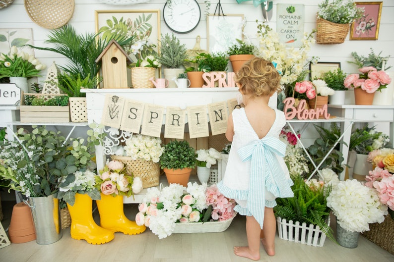Puede incluir: Una ni&ntilde;a peque&ntilde;a con un vestido blanco y un lazo azul se para frente a un estante blanco decorado con flores, plantas y una pancarta que dice "SPRING".