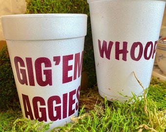 Op de afbeelding: Twee witte styroporbekers met donkerrode letters. De ene beker zegt "Gig 'Em Aggies" en de andere zegt "Whoop". De bekers staan op een bed van groen mos.