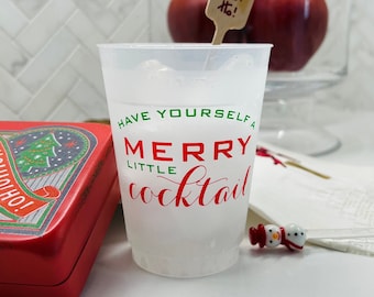 May include: A clear plastic cup with the text "Have Yourself a Merry Little Cocktail" in red and green. A red tin with a Christmas tree design is on the left. Two red apples and a wooden sign that says "Ho! Ho! Ho!" are in the background.