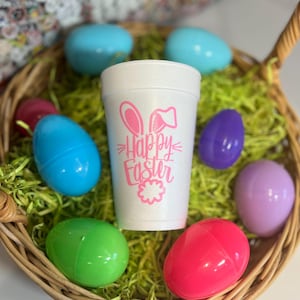 May include: A white foam cup with a pink "Happy Easter" design and bunny ears, surrounded by colorful plastic Easter eggs in a woven basket filled with green decorative grass.