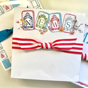 May include: A stack of white notepads featuring the word "NEWS" in colourful, decorative frames. The notepads are tied with a red and white striped ribbon. Other notepads with Asian-inspired designs are visible in the background.