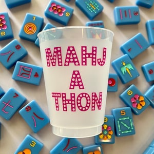 May include: A white plastic cup with the words "MAHJ A THON" printed in pink with white polka dots. The cup is surrounded by blue and white mahjong tiles.