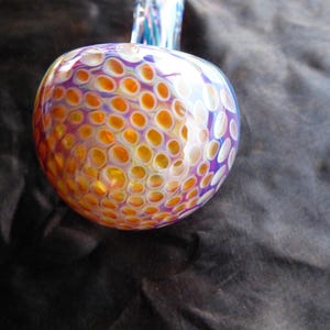 May include: A colorful, round glass art piece with a bulbous shape. The glass features a pattern of orange and yellow circles against a purple and white background. A clear glass stem extends from the top.