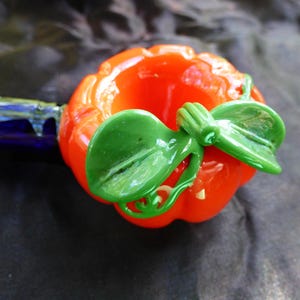 May include: A glass pumpkin pipe with a green leaf design. The pipe is orange and green and has a blue stem.