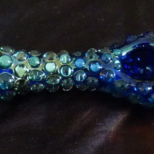 May include: A blue glass pipe with a textured surface. The pipe is decorated with small, round, blue and green glass beads.
