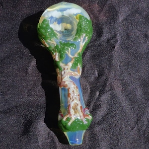 May include: A glass pipe with a tree design. The pipe is clear with a light blue tint and has a green tree painted on it. The tree has a brown trunk and green leaves. The pipe has a round bowl at the top and a long stem.