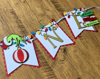 May include: A white and red banner with the word "ONE" in gold letters. The banner is decorated with green and red Christmas lights, a green Grinch hand, and a red and white striped ornament. The banner also includes signs with the words "Mt. Crumpit", "Whooville", and "Grinch's Lair".