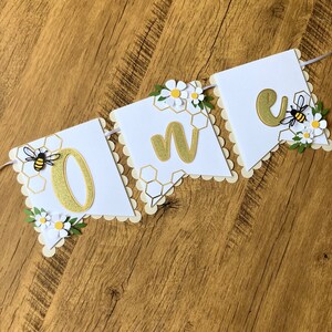 May include: White banner with gold glitter letters spelling "one" with a bee and honeycomb pattern on each flag. The banner is decorated with white paper flowers and green leaves.
