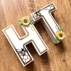 May include: White wooden letters "HT" decorated with gold trim, honeycomb pattern, and paper bees and sunflowers.