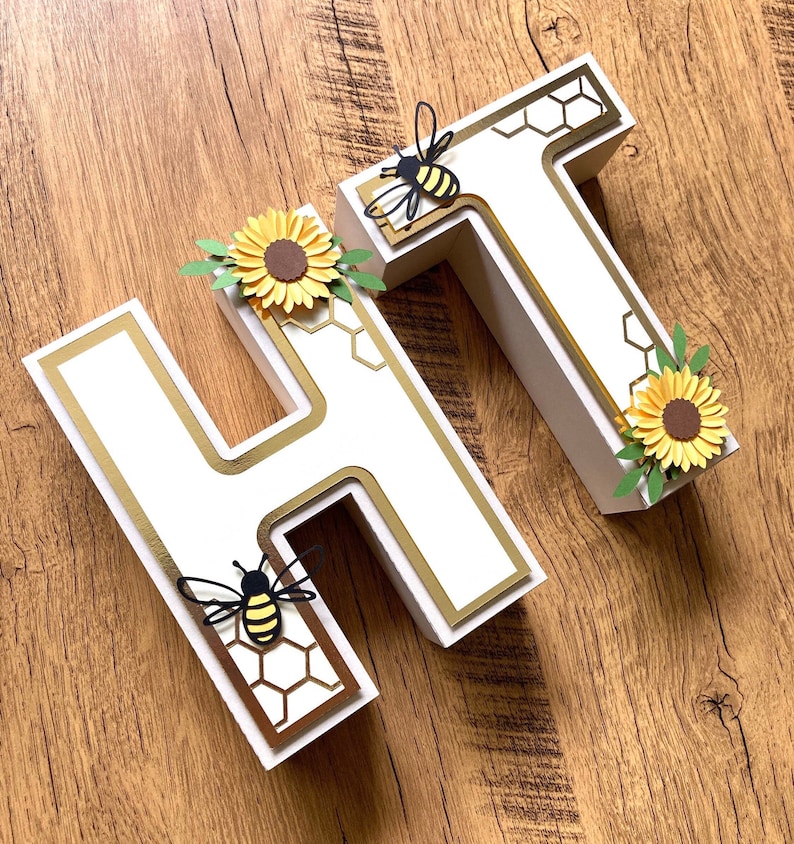 May include: White wooden letters "HT" decorated with gold trim, honeycomb pattern, and paper bees and sunflowers.