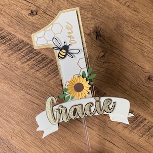 Puede incluir: Un adorno para pastel blanco con un número uno de purpurina dorada, una abeja, un girasol y hojas verdes. El adorno también tiene una pancarta blanca con el nombre "Gracie" en purpurina dorada.