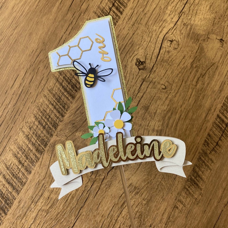 May include: A white cake topper with a gold glitter banner that says "Madeleine" and a number one with a bee and honeycomb design. The number one is decorated with white flowers.