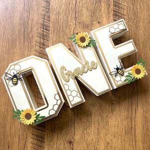 May include: White wooden letters spelling "ONE" with a honeycomb pattern and gold accents. The letters are decorated with sunflowers, leaves, and bees. The letter "N" has the name "Gracie" written in gold glitter.