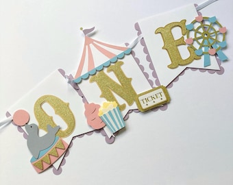 May include: A pink, blue, and gold banner with the word "ONE" in gold glitter letters. The banner also features a pink and blue circus tent, a popcorn bucket, a ticket, and a seal balancing on a ball. The banner is perfect for a first birthday party.