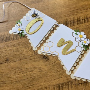 May include: White and gold banner with the word "One" in gold glitter letters. The banner is decorated with honeycomb patterns, white flowers, and a small bee.