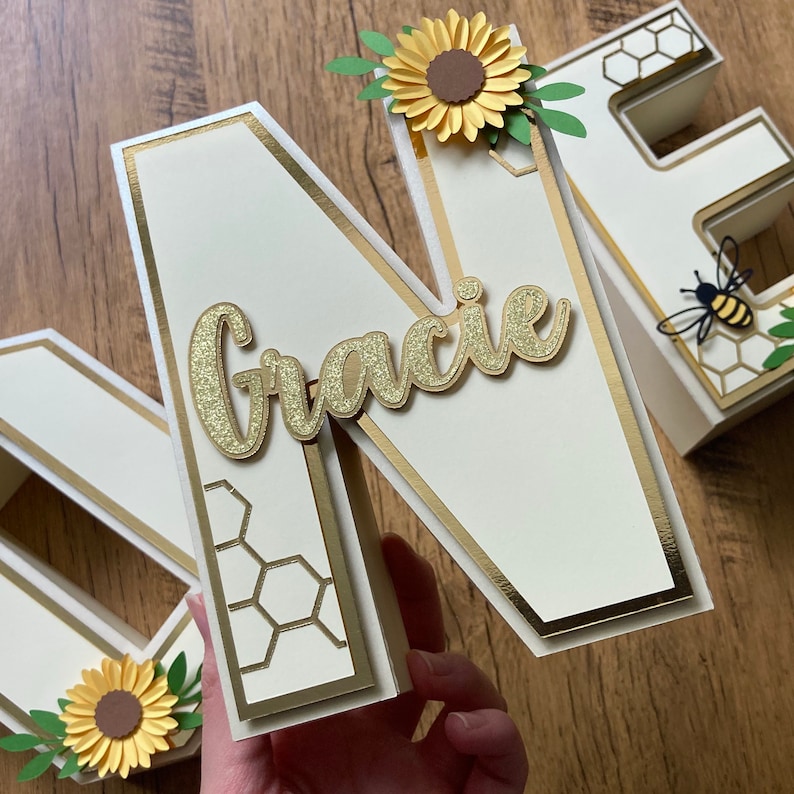 May include: White wooden letter "N" with a gold border and honeycomb pattern. The letter is decorated with a gold glitter name "Gracie" and a yellow paper flower with a brown centre.