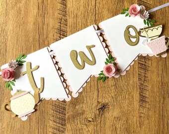 May include: A white banner with pink scalloped edges and gold glitter letters spelling "two". The banner is decorated with pink flowers, green leaves, and gold teapots and teacups.