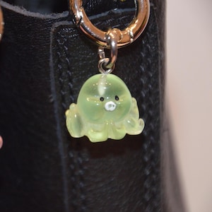 May include: A translucent green octopus charm with a small white mouth and black eyes, hanging from a silver-colored metal ring. The charm is attached to a black leather bag. The octopus has a cute, cartoonish design.
