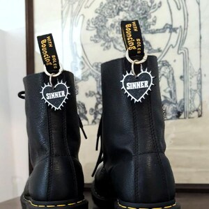 Könnte beinhalten: Paar schwarze Lederstiefel mit gelben Nähten und einer geriffelten Sohle. Jeder Stiefel hat ein Etikett mit der Aufschrift "Bouncing Soles" und einen herzförmigen Anhänger mit "SINNER".