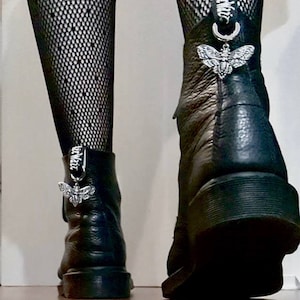 Może przedstawiać: Czarne skórzane buty z srebrnymi zawieszkami w kształcie ćmy przyczepionymi do kostki. Buty noszone są z czarnymi rajstopami w siatkę.