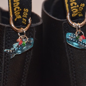 May include: Close-up of two black leather boots with decorative charms. Each charm is a blue whale shark with white spots and small red, green, and pink accents. The charms hang from silver hoops attached to the boot's pull tabs.