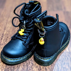 Puede incluir: Un par de botas de cuero negro para niños con dijes de patitos de goma amarillos. Las botas tienen cordones y un detalle de costura amarilla alrededor de la suela. El tamaño está etiquetado como UK 5, US 6, EU 20.