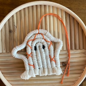 May include: A handmade macrame ghost ornament with a white body and orange accents. The ghost has black eyes and a fringed bottom. An orange string is attached for hanging. The ornament is displayed on a round, light brown wooden tray.