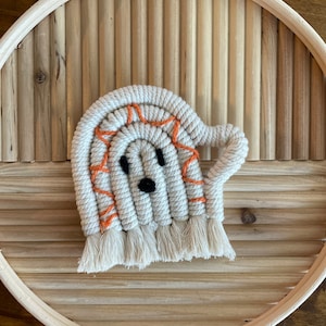 May include: A handmade macrame ghost coaster with a smiling face and orange accents. The ghost is made of off-white rope with a fringe at the bottom. The coaster is set on a round wooden tray with a natural finish.