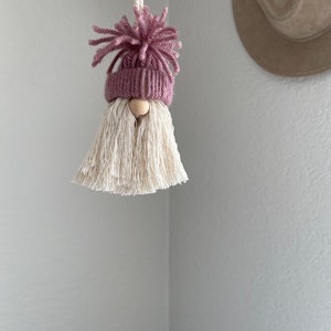 May include: A small pink and white gnome ornament with a white yarn beard and a pink yarn hat. The gnome is hanging from a white string.