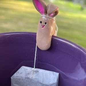 May include: A brown bunny tea bag holder with pink ears is hanging from a tea bag in a purple mug.