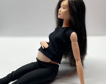 Puede incluir: Una muñeca embarazada con un top negro sin mangas y pantalones negros. La muñeca tiene el pelo largo y negro y está sentada sobre una superficie blanca.
