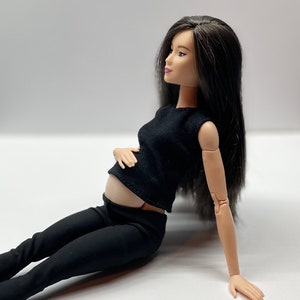 May include: A pregnant doll wearing a black sleeveless top and black trousers. The doll has long black hair and is sitting on a white surface.
