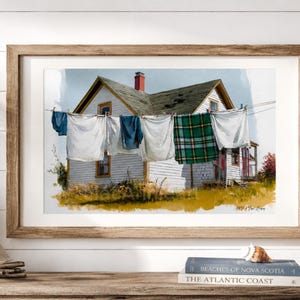 Peut inclure: Peinture à l'aquarelle d'une maison blanche avec une cheminée rouge et du linge suspendu à une corde à linge. Le linge comprend des draps blancs, un pantalon bleu et une couverture à carreaux verts. L'œuvre est encadrée dans un cadre en bois brun clair.