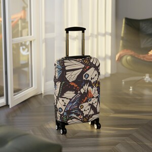 Peut inclure: Une valise à roulettes avec une poignée noire et une housse à motifs d'ailes de papillon en crème, bleu, orange et noir. La valise est posée sur un sol en bois.