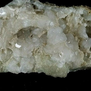 May include: A cluster of white, translucent crystals on a gray, textured rock. The crystals are clustered together in a random pattern, creating a visually interesting display.