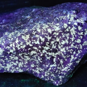 Könnte beinhalten: Ein großer, unregelmäßig geformter Stein mit einer violetten Basis und weißen Flecken. Der Stein wird mit UV-Licht beleuchtet, wodurch die weißen Flecken hell leuchten.