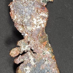 Puede incluir: Primer plano de un espécimen mineral multicolor de forma única. El mineral muestra una mezcla de texturas y colores, incluyendo tonos de marrón, naranja, gris y blanco. El espécimen se encuentra sobre un fondo oscuro.