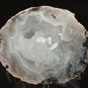 May include: A polished agate stone slice, showcasing concentric bands of gray, white, and translucent hues. The natural stone features a detailed pattern with dark brown and black edges, revealing its crystalline structure. A decorative mineral specimen.