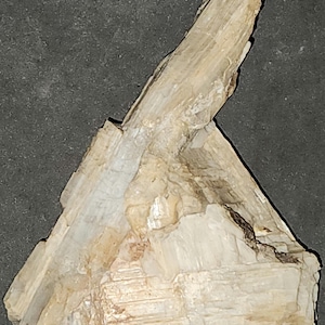 May include: A light beige mineral specimen with a layered, crystalline structure. The mineral has a rough, textured surface and a slightly translucent appearance. The specimen is set against a dark gray background.