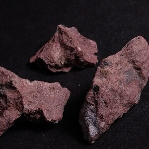 May include: Three rough, irregularly shaped pieces of red-brown rock on a black background.