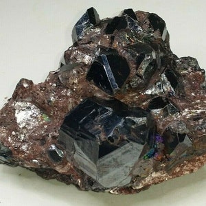 Puede incluir: Un grupo de cristales octaédricos de hematites negros e iridiscentes, incrustados en una matriz de roca marrón y gris.