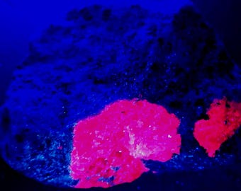 Op de afbeelding: Een close-up van een donkerblauwe steen met twee felroze gebieden. De steen heeft een ruw, getextureerd oppervlak. De roze gebieden zijn gemaakt van kleinere kristallen.