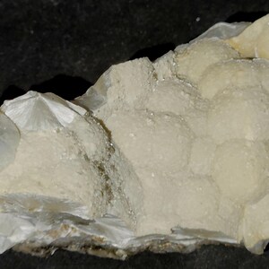 May include: A close-up of a natural mineral specimen. The off-white stone features crystalline structures and a bubbly texture. The specimen has a rough, uneven surface with a few clear, crystal-like formations.