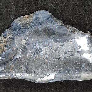 May include: A piece of flint rock with a rough, irregular shape. The stone has a mix of gray, silver, and brown tones, with a slightly reflective surface. The texture appears coarse and natural, suggesting it's a raw, unprocessed material.