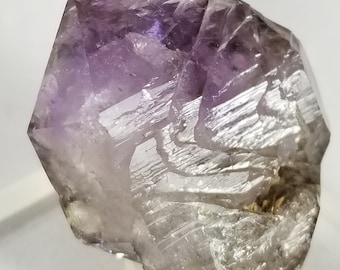 May include: A large, clear amethyst crystal with a purple hue and a unique, swirling pattern. The crystal is faceted and has a pointed tip.